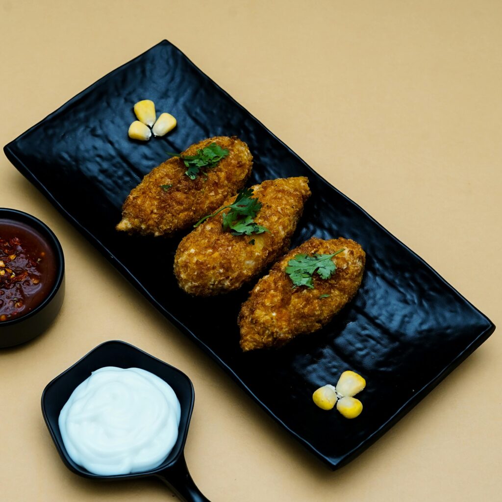 Appetizing corn croquettes served with red and white dip on a black plate.